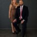 A woman and man in formal attire pose confidently against a dark background.