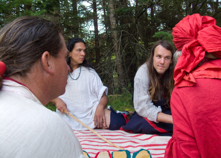 Four people in historic clothing facing each other around a striped blanket with leather pieces on it.