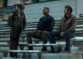 Three men sitting on stadium bleachers, one wearing a cowboy hat.