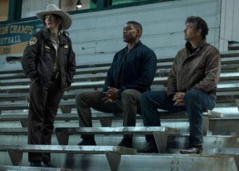 Three men sitting on stadium bleachers, one wearing a cowboy hat.