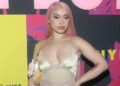 Ice Spice with long pink hair poses in a pale outfit at a colorful event backdrop.
