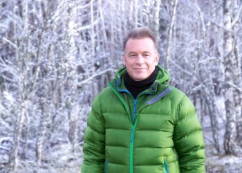 An individual dressed in a green puffer jacket with a black inner shirt, standing confidently in front of a backdrop of frost-covered trees.