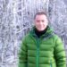 An individual dressed in a green puffer jacket with a black inner shirt, standing confidently in front of a backdrop of frost-covered trees.