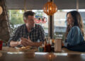 Josh Duhamel and Minka Kelly sit together at a diner counter in a scene from Ransom Canyon. Duhamel, wearing a blue plaid button-down shirt, looks toward Kelly with his hands folded beside a plate of pancakes. Kelly, dressed in a loose blue overshirt over a dark top, smiles back at him. Condiment bottles, a napkin dispenser and a coffee cup sit on the counter between them, while large windows and warm pendant lighting create a cozy, small-town diner atmosphere.