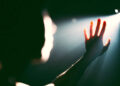A hand reaching towards a bright light in a dark setting, with the figure's silhouette partially visible.