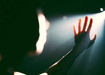 A hand reaching towards a bright light in a dark setting, with the figure's silhouette partially visible.