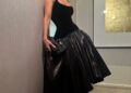 Woman in elegant black dress with a fitted top and voluminous skirt, posing against a wall in a stylish interior setting.
