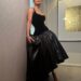 Woman in elegant black dress with a fitted top and voluminous skirt, posing against a wall in a stylish interior setting.