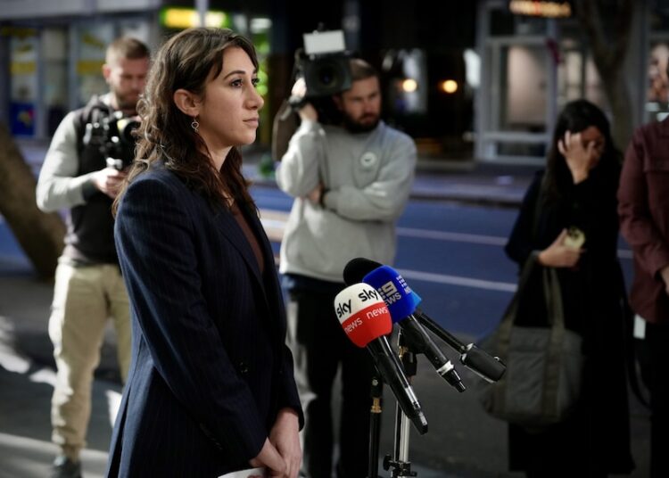 Ms Schwartz in a black blazer looking serious as she speaks into microphones outside a building.