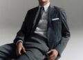 A young man in a stylish gray suit sitting on a wooden chair against a neutral background.