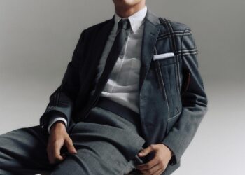 A young man in a stylish gray suit sitting on a wooden chair against a neutral background.