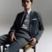 A young man in a stylish gray suit sitting on a wooden chair against a neutral background.