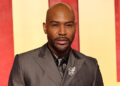 Karamo Brown in a gray suit with a brooch stands in front of a red and yellow background.