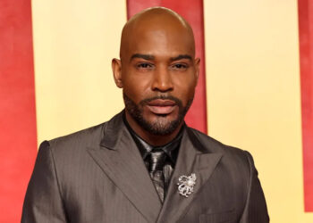 Karamo Brown in a gray suit with a brooch stands in front of a red and yellow background.