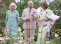 King Charles and Queen Camilla with garden designer Catherine MacDonald are in a garden. Green-leaved trees form the dense backdrop with yellow and purple flowers in the foreground. A pale lemon-coloured hydrangea can be seen too. The Queen is wearing a green-patterned midi dress, MacDonald is wearing a yellow midi dress with a flower print and King Charles is wearing a beige linen suit and is carrying a brown walking stick.