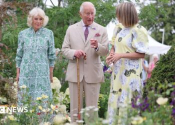 King Charles and Queen Camilla with garden designer Catherine MacDonald are in a garden. Green-leaved trees form the dense backdrop with yellow and purple flowers in the foreground. A pale lemon-coloured hydrangea can be seen too. The Queen is wearing a green-patterned midi dress, MacDonald is wearing a yellow midi dress with a flower print and King Charles is wearing a beige linen suit and is carrying a brown walking stick.