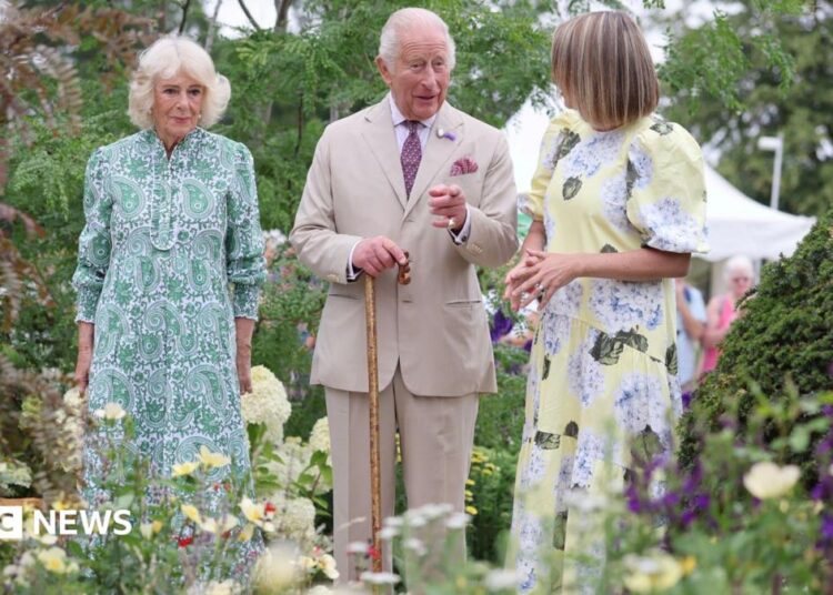 King Charles and Queen Camilla with garden designer Catherine MacDonald are in a garden. Green-leaved trees form the dense backdrop with yellow and purple flowers in the foreground. A pale lemon-coloured hydrangea can be seen too. The Queen is wearing a green-patterned midi dress, MacDonald is wearing a yellow midi dress with a flower print and King Charles is wearing a beige linen suit and is carrying a brown walking stick.