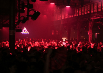A lively crowd gathered in a dimly lit venue with red lighting and a neon sign in the background.