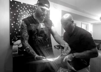 Maxx Crosby, wearing a chain with an "E" pendant, holds a bright, glowing object in a black and white image.