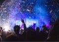 Silhouetted crowd at an outdoor gig at night, the stage is lit with blue light and confetti falls.