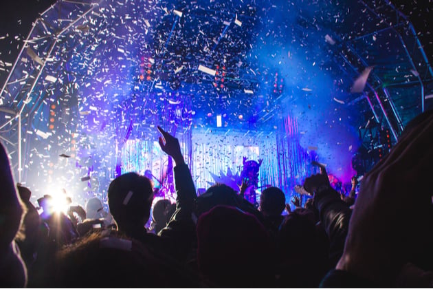 Silhouetted crowd at an outdoor gig at night, the stage is lit with blue light and confetti falls.