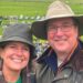 Alice Dearing and her husband John stood next to each other having their picture taken outside. They are dressed in coats and hats each and are looking directly at the camera and smiling. Alice is on the left and John is on the right. They are at an event with crowds of people in the background.