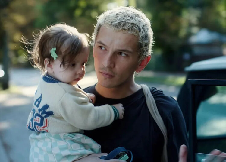 Filip Raczyński in a scene from "Proud." Young man with short bleached hair carries a toddler girl while standing next to a car.