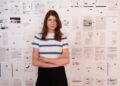 A woman in a white T-shirt with blue stripes stands with her ams crossed in front of a collage of text and documents.