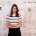 A woman in a white T-shirt with blue stripes stands with her ams crossed in front of a collage of text and documents.