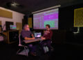A faculty member and student sit together in an audio production studio reviewing a multitrack recording project displayed on a laptop and large projection screen, discussing audio editing and sound production techniques.