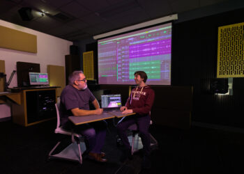 A faculty member and student sit together in an audio production studio reviewing a multitrack recording project displayed on a laptop and large projection screen, discussing audio editing and sound production techniques.