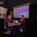 A faculty member and student sit together in an audio production studio reviewing a multitrack recording project displayed on a laptop and large projection screen, discussing audio editing and sound production techniques.