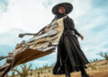 Still from "Blades of the Guardians." Low-angle image of a person in a long black robe and wide-brimmed hat dragging a large bundle of weapons across a barren landscape under a cloudy sky.