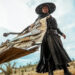Still from "Blades of the Guardians." Low-angle image of a person in a long black robe and wide-brimmed hat dragging a large bundle of weapons across a barren landscape under a cloudy sky.