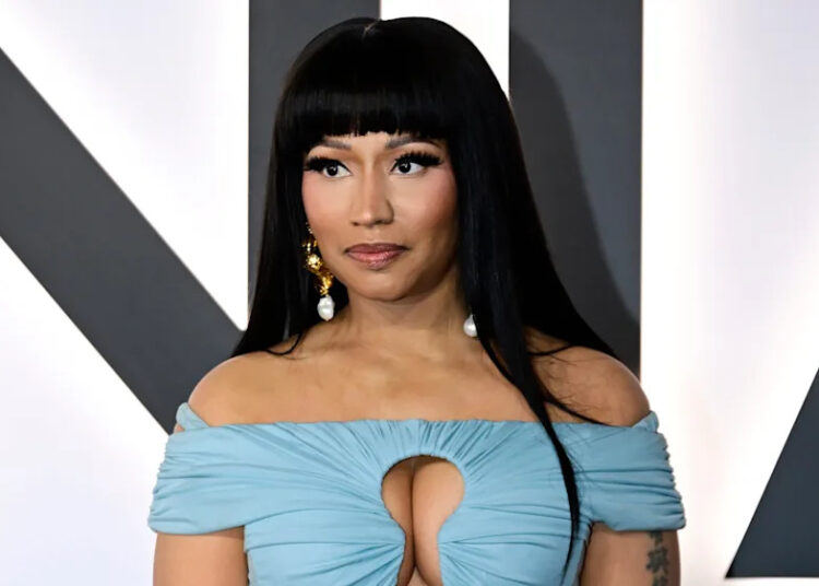 Nicki Minaj in a light blue off-shoulder dress with gold bracelets, standing against a neutral background.