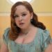 A young woman with red hair wears a blue dress and jewelry, looking thoughtfully at the camera.
