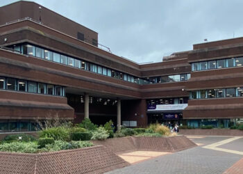 A building of at least three storeys, with the image showing one part of the building on the left at right angles to another part of the building on the right. An area for pedestrians is in the foreground.