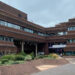 A building of at least three storeys, with the image showing one part of the building on the left at right angles to another part of the building on the right. An area for pedestrians is in the foreground.