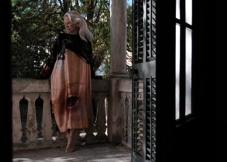A woman stands on a balcony wearing a long artistic dress with a striking design, surrounded by greenery and natural light.