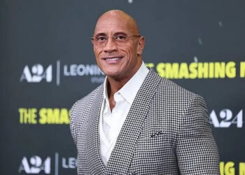 A person in a stylish checkered suit smiles at a media event backdrop featuring the text "A24" and "The Smashing."
