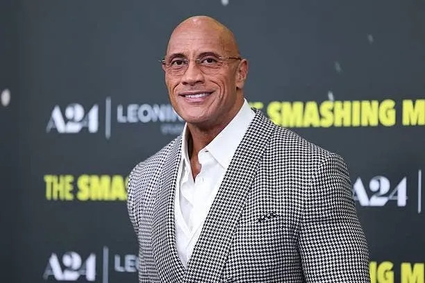 A person in a stylish checkered suit smiles at a media event backdrop featuring the text "A24" and "The Smashing."
