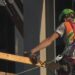 A close-up shot of the back of a construction worker holding a drill while wearing a high-visibility vest.