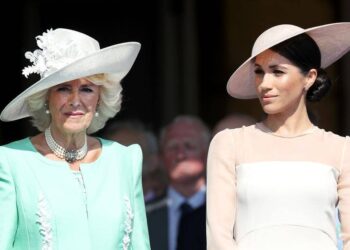 Meghan, Duchess of Sussex, in a white dress and hat, stands next to queen camilla in a green dress and white hat.