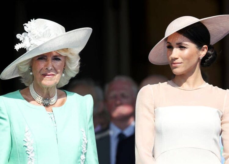 Meghan, Duchess of Sussex, in a white dress and hat, stands next to queen camilla in a green dress and white hat.