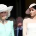 Meghan, Duchess of Sussex, in a white dress and hat, stands next to queen camilla in a green dress and white hat.