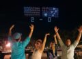 Four people cheering outdoors at night with two drone-formed flags and "2 - 0" displayed in the sky above them. Trees and city lights are visible in the background.