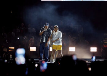 Jay-Z and Pharrell on stage, one in a cap and jeans, the other in a hat and yellow shorts, with a crowd and bright lights in the background. This is night two of Jay-Z's Yankee Stadium series in the Bronx. This night he played The Blueprint.