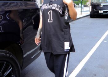 A person wearing a sports jersey and pants walks beside a black vehicle in an urban setting.