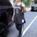A person wearing a sports jersey and pants walks beside a black vehicle in an urban setting.