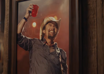 Man in a cowboy hat cheers with a red cup in a framed portrait against a warm, softly lit background.